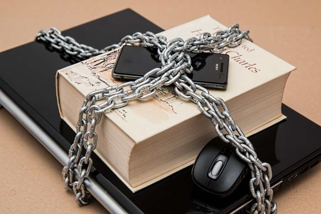 A laptop, mouse, cell phone, and book are bound together by heavy chain.